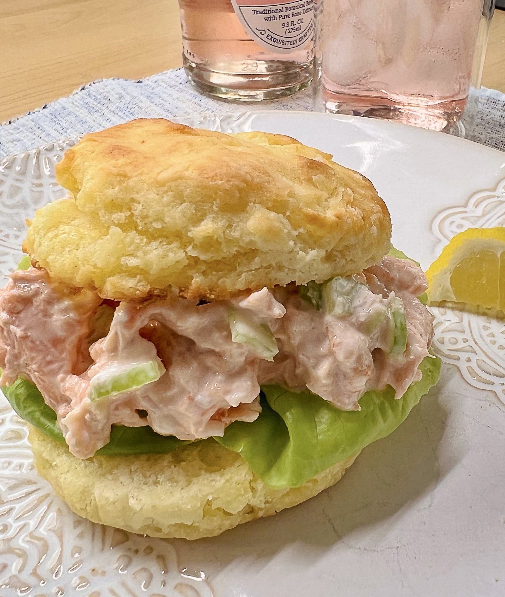 salmon salad on a buttermilk biscuit with bibb lettuce sitting on a scalloped decorated cream-colored plate, wedge of lemon to the right, pink lemonade over ice in a tall clear glass.