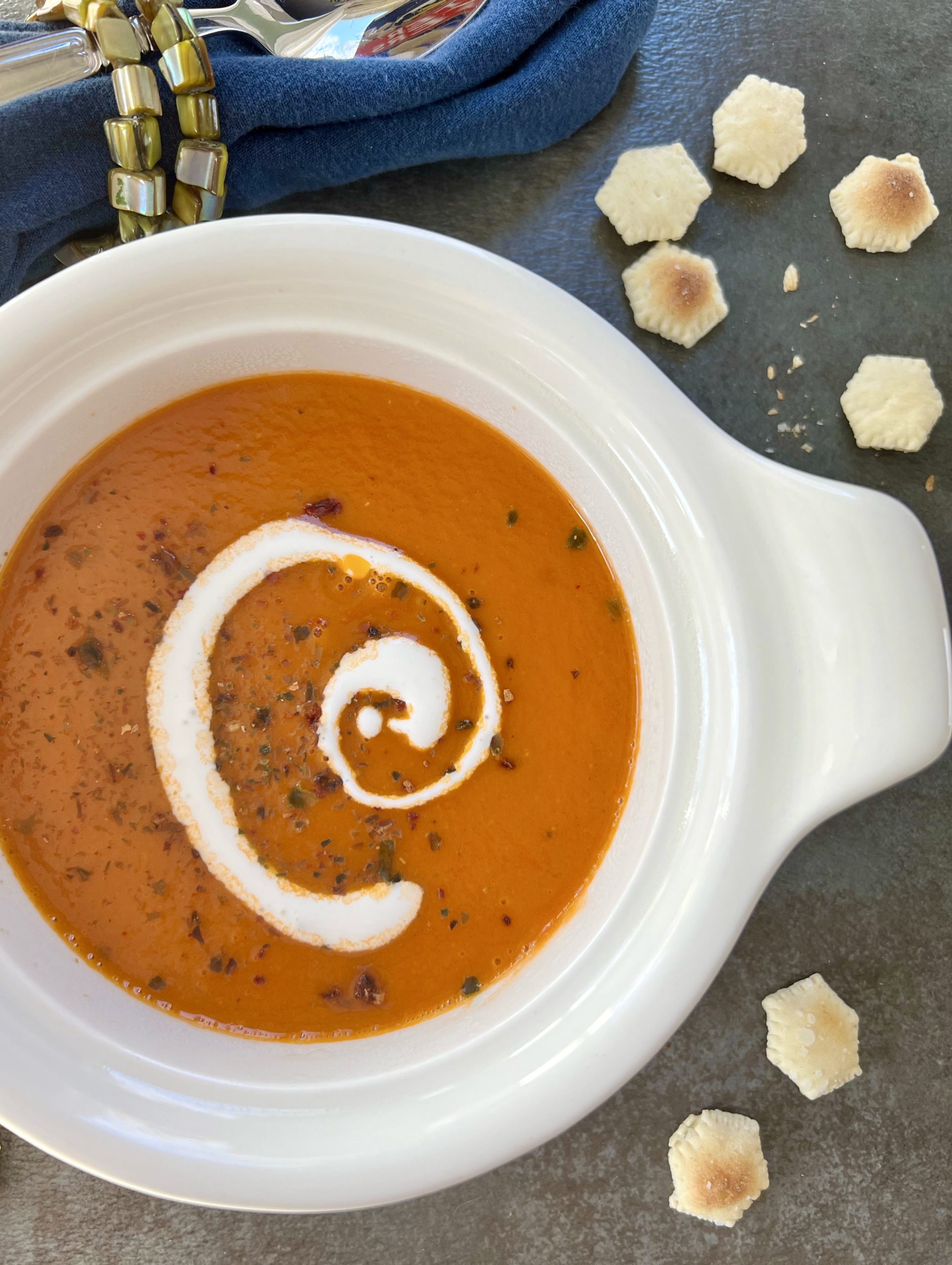 Overhead shot of creamy Simple Homemade Tomato Soup in a white porcelain crock surrounded by oyster crackers. A swirl of Mexican Crema swirl tops the soup along with spicy seaweed flakes. Soup bowl sits on a navy blue background with a blue napkin and spoon peaking off the top left of the image.