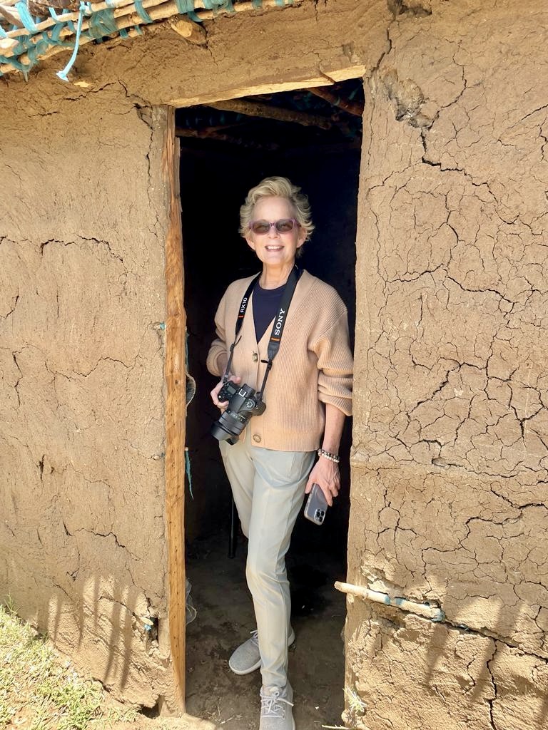 maureen c berry stands in a dorrway of an masai mara hut wearing a camera around her neck and holding a iphone in her hand wearing a beighe sweater, olive green slacks and grey sneakers