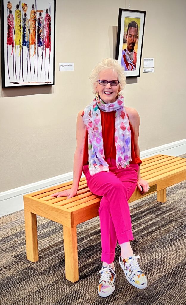 maureen c. berry, sits on a beige wood bench in front of two pieces of art, acrylic amasai mara men aand a photograph on a beige wall. she wear bright pink pants, red blouse and pastel colored neck scarf.